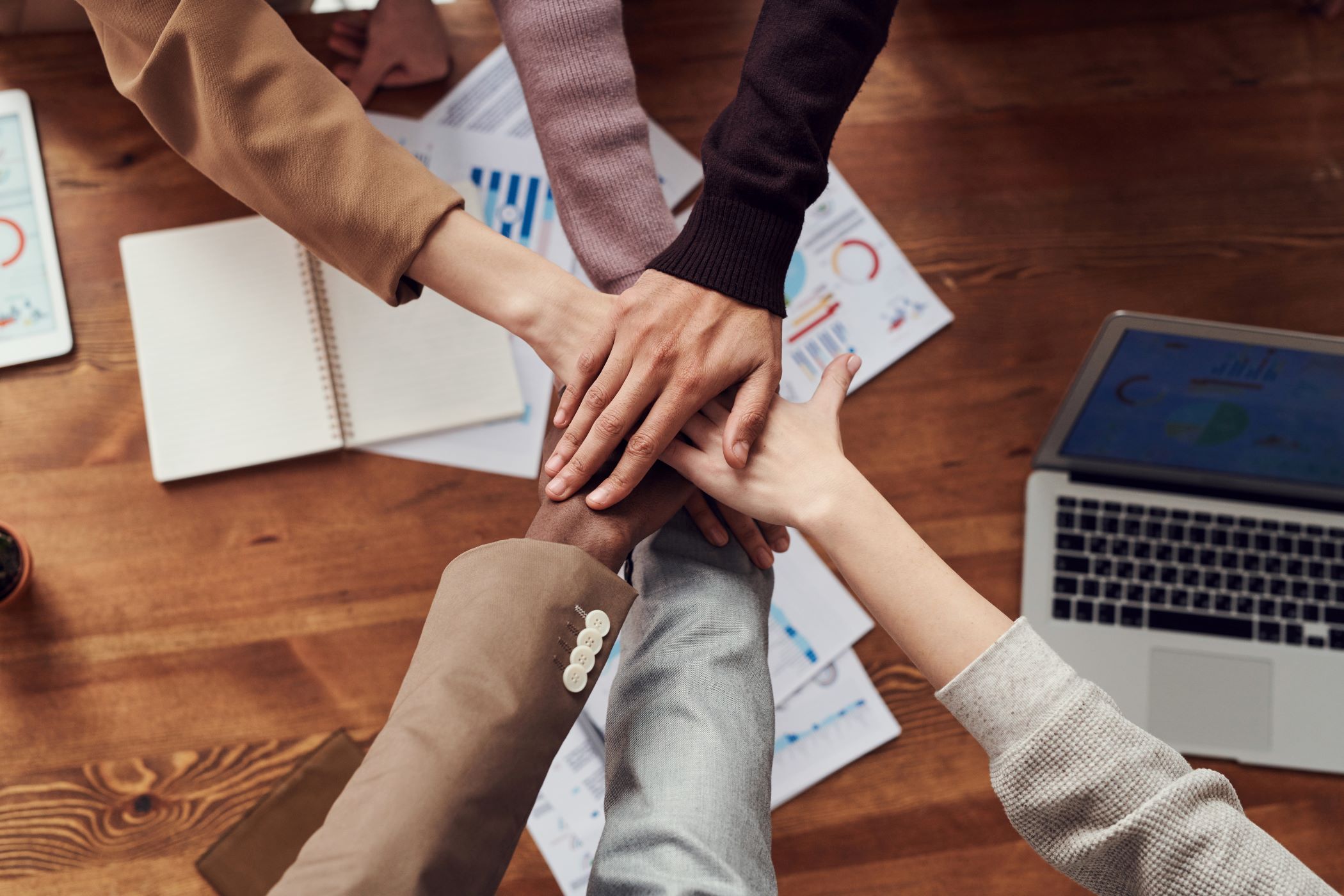 Coworkers layer hands over a wooden conference table with marketing analytics and open laptops.